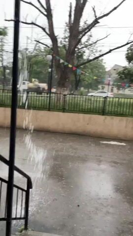 The video captures a street scene during a heavy downpour. The ground is wet and reflects the light, with raindrops visibly hitting the surface. A black railing and a concrete barrier are in the foreground. In the background, there are trees, a building, and cars on a road. Some colorful flags are strung between trees. A lamppost with a light fixture is visible on the left side of the screen, and water is gushing down a pole near a set of stairs on the right. The overall atmosphere is one of intense rain.