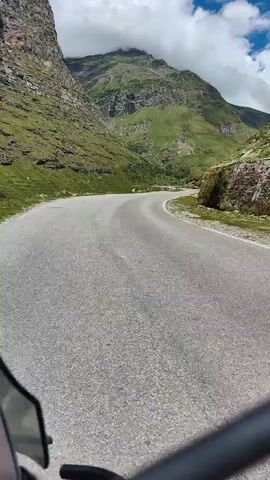 The video shows a view from a motorcycle on a paved road winding through a mountainous landscape. The road is asphalt and appears to be in good condition, with some cracks visible. The surrounding environment is characterized by steep, green, grassy mountainsides. The sky is partly cloudy with patches of blue sky. The camera perspective suggests the motorcycle is in motion, traveling along the road.