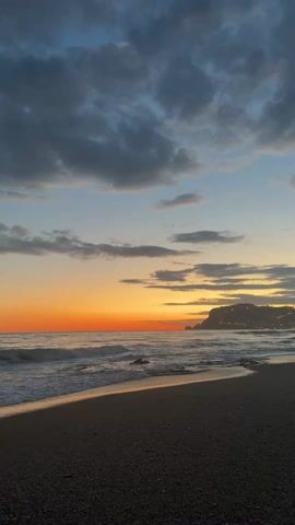 The video shows a beach at sunset. The sky is a mix of dark blue, gray, and orange. There are clouds scattered across the sky. The ocean is visible, with small waves rolling in towards the shore. The beach is made of dark sand and small pebbles. In the distance, there is a land mass with lights on it. The scene is still and quiet.