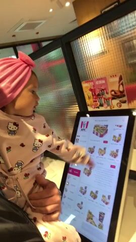 A man is standing in a KFC restaurant holding a small child, who is wearing a pink outfit with Hello Kitty designs and a pink turban. The child is using the touch screen ordering kiosk at the restaurant, tapping different menu options with her finger. The man is smiling and looking at the camera. There is a promotional poster behind the ordering kiosk with the words "BUBBLE TEA ICE CREAM CHURROS".