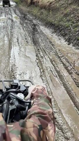 A person wearing camouflage clothing drives an all-terrain vehicle through a muddy, rutted road. The person is wearing black boots and the vehicle is traveling behind another ATV. The camera is mounted on the ATV to provide a first-person perspective of the journey. The path is muddy and wet and surrounded by trees. The muddy road is visible ahead.