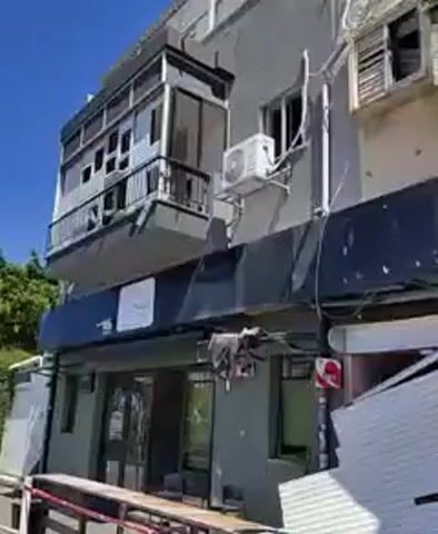 The video shows damage to a building, seemingly a store, with a metal roll-down security door severely damaged and bent inwards. The door has multiple puncture holes, suggesting impact damage.  The building is light-colored, with a black awning and a balcony with broken glass. Red and white barrier tape stretches across the front of the store. The environment appears to be urban, and the lighting is bright.