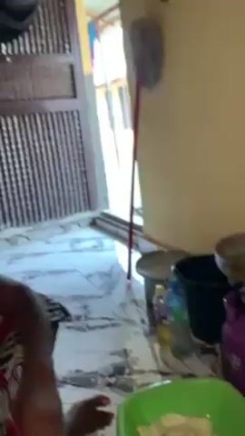 A woman wearing a patterned fabric wrap is shown sitting on a marble floor, preparing food in a green bowl. Her hair is braided back tightly. She appears to be in a kitchen area. To her right is a black bucket, a water bottle, a pink scale, and a pot on a stovetop. Behind her is a metal screen door.  The walls are painted a light color.