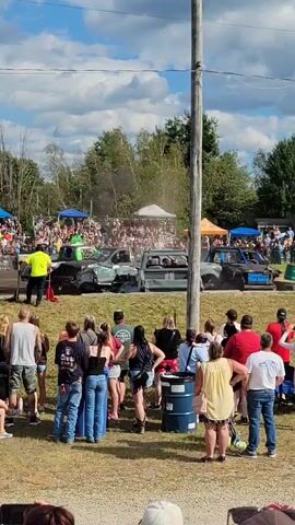 A demolition derby event is underway with several heavily damaged cars in the arena. A crowd of spectators is gathered, some seated in bleachers in the background and others standing closer to the action. In the foreground, a group of onlookers stands behind a low fence. The sky is blue with scattered clouds. There are utility poles and trees visible in the background. A few individuals in safety vests and helmets appear to be officials or workers attending to the vehicles. The atmosphere seems lively, with dust or smoke rising from the derby area.