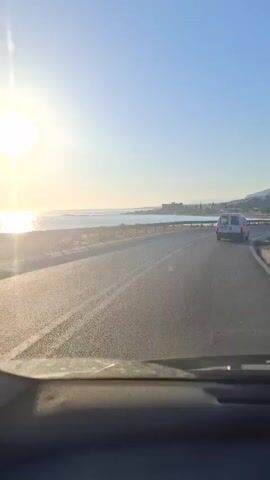 The video shows a view from inside a car driving along a road next to the sea. The sun is shining brightly and low in the sky, creating a glare. There is a white van driving in front of the car. The road curves to the right, and the sea is on the left. There are buildings visible in the distance along the coastline.