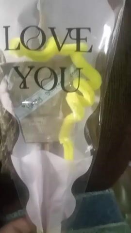 The video captures a close-up view of a bouquet wrapped in clear cellophane. The bouquet features a tag with the word "YOU" printed on it, and what appears to be a handwritten inscription below. Inside the cellophane, there are light pink and white decorative papers, and a bright yellow curly ribbon. At the bottom of the bouquet, a yellow ribbon bow is tied, with a small pink embellishment in the center and two small beige ribbon tendrils extending downwards. A hand is visible in the lower left corner of the frame, holding the base of the bouquet. The background is blurry, suggesting a dimly lit indoor setting, possibly a table covered with a dark blue textured cloth.