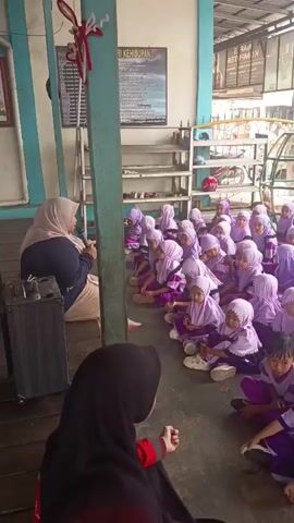 The video shows a group of young children, most wearing purple hijabs and purple and white uniforms, sitting on the floor in rows. An adult woman, also wearing a hijab and a blue and tan outfit, is sitting on a raised wooden platform to the left of the frame and appears to be speaking to the children, holding a small black object, possibly a microphone. Another adult woman, wearing a dark hijab and a patterned top, stands near a gate in the background. The environment appears to be outdoors, with a covered ceiling overhead and buildings and a playground structure visible in the background. The children are attentively looking towards the speaker.