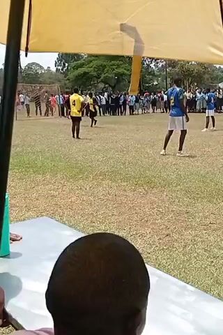 The video shows a game of dodgeball taking place on a grassy field. Players are wearing different colored shirts, with some in yellow and others in blue. Spectators stand on the sidelines, watching the game. The camera is positioned under a yellow tent, providing a partial view of the field and the action. One person is sitting in front of the camera. The background includes trees and a clear sky.