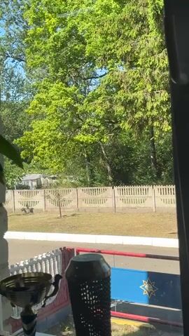 The video captures a scene viewed through a window. In the foreground, on the windowsill, are two objects: a trophy with blue and white accents and a dark, cylindrical object with a textured surface. Beyond the window, a light-colored fence runs horizontally across the frame, with a patterned design. Behind the fence, lush green trees dominate the background, with some of the trees appearing to be evergreens. The sky above is a clear blue, suggesting fair weather.

The main event unfolding is the approach of a bright red fire truck. The truck has white and blue accents and appears to be moving from left to right across the frame. On the side of the truck, the word "СЛОНІМ" (Slonim) is visible in white lettering, and there is a number "1" in a blue rectangle. There is also a symbol that resembles a star within a circle. The truck's headlights are on, indicating it might be in use.

The presence of the fire truck suggests an emergency response or a routine activity at a fire station. The overall atmosphere seems calm, with the bright daylight illuminating the scene. The location is confirmed to be Slonim, Belarus, by the text on the fire truck. The view through the window and the objects on the windowsill provide a domestic or office perspective on the event outside.