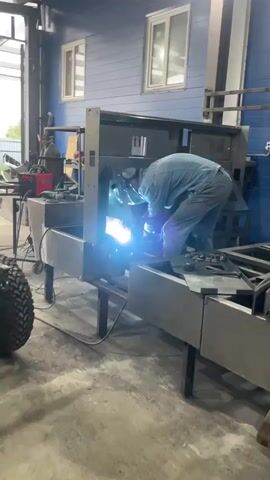 A person wearing a denim jumpsuit and a welding mask is actively welding a large metal structure. Sparks are flying from the point of welding, illuminating the immediate area. The person is bent over the structure, focused on the task. The structure itself appears to be part of a larger industrial or commercial fabrication, with several metal components and supports visible. The environment is a workshop or factory setting, with industrial-style walls and windows in the background. Other equipment, including what looks like a welding machine and a tire, are present in the vicinity. The lighting suggests an indoor space with artificial light sources.