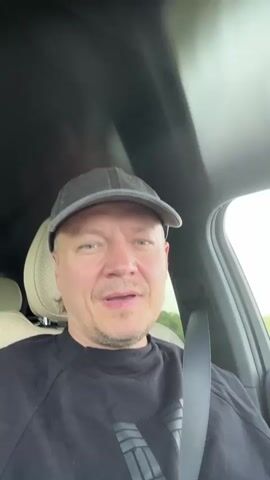 A man wearing a black baseball cap and a black t-shirt with a grey design is sitting in the driver's seat of a car. He is looking at the camera and smiling. The interior of the car is visible, including the headrest of the passenger seat and the seatbelt. The car appears to be moving, as indicated by the blurred background outside the window and the sound of wind. The man touches his cap and then his head. The overall scene is casual and appears to be a personal video recorded while driving.