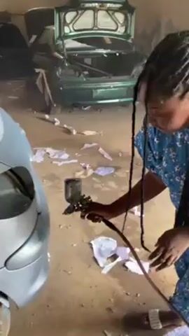 A woman is shown in a garage painting a car. She is wearing a blue dress with a floral print and has her hair in braids. She is holding a paint sprayer with a red hose attached. The car is light blue and partially obscured by the woman. In the background, there is a green car with its trunk open. There are pieces of white paper scattered on the floor. The garage appears to be dusty.
