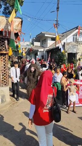 A street parade is happening on a sunny day. People in costume and regular clothes are walking along a paved street lined with houses. A person in a furry monster costume with a mask is prominent. A small child wearing a loincloth and headwrap dances. People in the crowd wave and take photos with their phones. Banners and flags are displayed along the route. Some of the parade participants are in red tops, others wear traditional clothing.