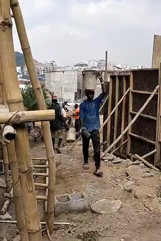 The video shows several men working on a construction site. They are carrying buckets of material, some on their heads, up a wooden ladder. The construction site is located in an urban area with buildings visible in the background. The weather appears to be overcast. There is a bamboo scaffolding on the left side of the frame.  The workers are wearing casual clothes, and some have head coverings. The ground is dirt and gravel.