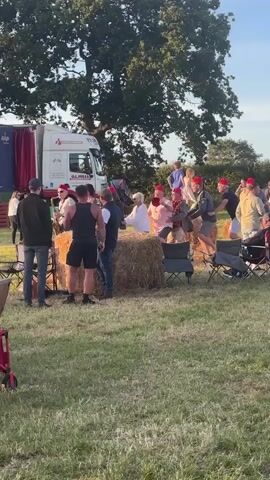 A crowd of people are gathered outdoors on a grassy field. Some are wearing red hats. A truck with a trailer is parked in the background, with a stage set up in front of it. Several bales of hay are scattered around the area, serving as seating. Some individuals appear to be dancing. The overall atmosphere suggests an outdoor event or festival.