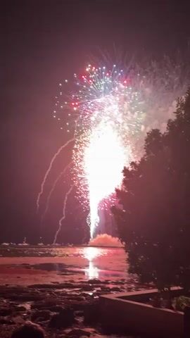The video shows a fireworks display over a body of water at night. The fireworks are exploding in various colors, including red, pink, white, green, and blue. Some fireworks are shaped like bursts, while others are trails of light. The reflection of the fireworks can be seen on the surface of the water. In the foreground, there is a rocky shoreline with some vegetation on the right side of the frame. There are also a few boats visible in the distance on the water. The sky is dark, creating a contrast with the bright explosions of the fireworks. There appear to be a few people in the lower left corner of the frame, possibly watching the display. The overall scene is festive and visually striking, capturing the excitement of a fireworks show.