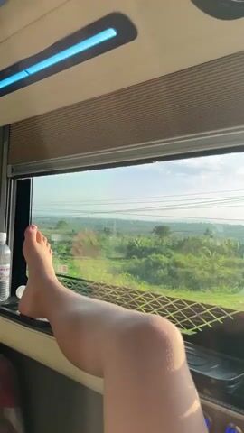 The video opens with a close-up of a person's bare legs and feet, propped up against the window ledge inside what appears to be a train or bus. A clear plastic water bottle is visible next to their feet. The interior of the vehicle shows a window with a pulled-up blind and an overhead blue light fixture.

The camera then pans and zooms out to capture the view outside the window. The vehicle is moving, showcasing a landscape of green fields, possibly agricultural land, under a bright, clear sky. Numerous overhead power lines traverse the frame horizontally. In the distance, there are scattered trees, some distant buildings, and faint hills or mountains. The light suggests it is daytime, likely a sunny morning or afternoon. The scene outside transitions from open fields to areas with more vegetation and poles, indicating a journey through a rural or semi-rural area. The visible elements confirm the location context of Thôn 4, Vietnam. No discernible text is visible in the video.