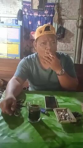 A man wearing a green t-shirt and a yellow baseball cap sits at a green table. He is smoking a cigarette and exhaling smoke. On the table in front of him is a glass of dark liquid, a lighter, a smartphone, and an ashtray filled with cigarette butts. The background appears to be an indoor setting with a woven wall.