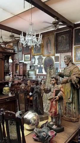 The video shows the interior of a room filled with antique furniture, artwork, and statues. Walls are completely covered with framed pictures, primarily portraits and religious iconography, like an image of the Virgin of Guadalupe. A crystal chandelier hangs from the ceiling, which also features dark wooden beams and several ceiling fans. In the foreground, there's a table displaying several wooden statues of religious figures, along with a mirrored gazing ball and a decorative animal mask. Other furniture pieces, like cabinets, chairs, and a china cabinet, are placed throughout the room. Overall, the scene creates a cluttered but curated impression of an antique shop or a collector's storage.
