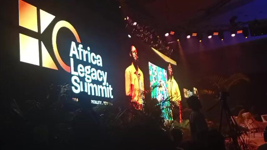 The scene is a large, dimly lit conference hall. A large screen to the left displays the logo for the "Africa Legacy Summit", and a screen to the right shows two individuals in orange shirts, followed by a photo of a group of people. There are spotlights and stage lighting illuminating the screens and stage area. In the foreground, there is a camera on a tripod and some decorative plants. There are also silhouettes of people in the audience. The focus is on the screens and the event being presented. The overall impression is that of a professional conference or summit.