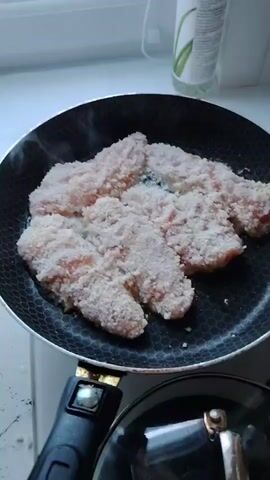 In a close-up shot, several breaded chicken tenders are sizzling in a non-stick frying pan on a stovetop. The pan has a hexagonal pattern on its surface and is positioned over a burner, indicated by the steam rising from the chicken. A black pot lid with a silver handle is partially visible in the foreground, positioned below the frying pan. The background is blurred, but hints of a white countertop and a bottle with green on it can be seen. The lighting suggests an indoor setting, possibly a kitchen. The overall impression is one of cooking food.