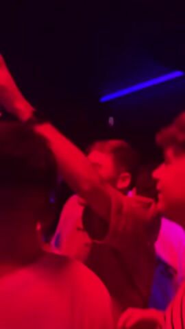 The video shows a dimly lit club with red lighting. In the foreground, a person is visible with their arm raised, holding a glass. Other people are present in the background, some blurry, suggesting movement or low light. The environment appears to be a crowded venue, likely a nightclub or bar, with a discoball visible in the upper portion of the frame. The lighting creates a vibrant, energetic atmosphere.