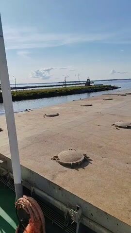 The video shows a flat, concrete surface with several circular metal hatches. The surface appears to be the deck of a vessel or a pier. In the background, a calm body of water is visible, with a narrow strip of land and a lighthouse on the horizon. The sky is blue with a few scattered clouds. The lighting suggests it is daytime.