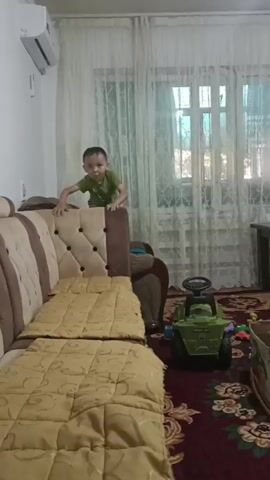 A young boy in a green shirt and shorts is captured mid-air, appearing to jump off the armrest of a large, upholstered sofa. The sofa is covered with a beige and brown patterned throw blanket. To the right of the sofa, a green toy tractor is parked on a dark red carpet with an intricate floral pattern. The room features sheer white curtains covering a window. In the background, a dark wooden table with a bag on it is visible. Further to the right, a girl in a white dress with floral patterns is sitting on the floor, surrounded by a variety of colorful building blocks and toy pieces. Two chess boards are placed on the carpet in front of her. A large yellow dump truck toy is also present. The lighting suggests an indoor daytime environment.