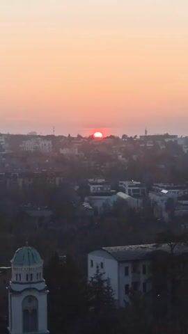The video shows a cityscape at sunrise or sunset. The sky is a gradient of orange and peach, with the sun appearing as a red disk just above the horizon. The city below is a mix of buildings and trees, with a church steeple visible in the foreground. A jet stream can be seen briefly in the upper part of the shot.