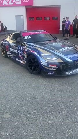 A blue and gold drift car with the number 19 on the windshield is parked on asphalt in front of a building with red garage doors and a sign that says "NR СЕРВИС". Several people, some wearing yellow shirts, stand around the car. A person in a white helmet is visible in the driver's seat. The car has various sponsor logos on its side, including "TAKAYAMA" and "LINGLONG". The camera pans across the scene.