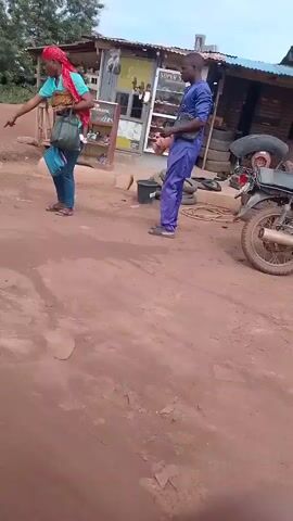 The video opens with a shot of a dusty, unpaved road. In the background, a few people are visible near a small open-air shop. A man in a purple outfit stands near the shop, facing left. To his right, a woman with a red headscarf and a blue shirt walks away from the camera, carrying a bag. A pink motorcycle is parked in front of the shop. The camera then pans to the right, showing more of the shop and the road. A silver minivan is parked further down the road in the distance. The camera continues to pan, and the video ends with a close-up of what appears to be a wooden pole or stick in the foreground. The environment suggests a rural or semi-urban setting.