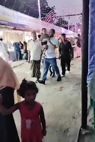 The video shows a bustling outdoor market or fair in what appears to be Charfasson, Bangladesh, likely during the evening or night. The scene is illuminated by numerous string lights and decorated arches, creating a festive atmosphere. Many people, including men, women, and children, are seen walking along a pathway lined with temporary stalls. Some women are dressed in traditional Islamic attire, including hijabs and niqabs. Men are also present, some carrying young children on their shoulders or by hand. The ground is a mix of dirt and possibly concrete, with some visible debris or trash. People appear to be casually moving through the area, browsing, and socializing. The overall impression is that of a lively community gathering or market event. No legible text is visible in the video.