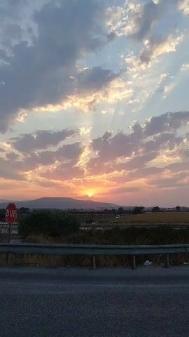 A vertical shot pans across a sunset sky. The sun, a bright orange orb, is low on the horizon, peeking over a line of distant mountains. Rays of light beam outwards from the sun through scattered clouds, which are tinged with pink and orange hues. In the foreground, a road is visible, with a metal guardrail separating it from a grassy area. A red and white stop sign with the Turkish word "DUR" is planted near the guardrail. Some vegetation, including green bushes and dry grasses, are present in the mid-ground. A white vehicle is visible on a road in the distance. The sky transitions from bright orange and yellow near the sun to a pale blue at the top of the frame.