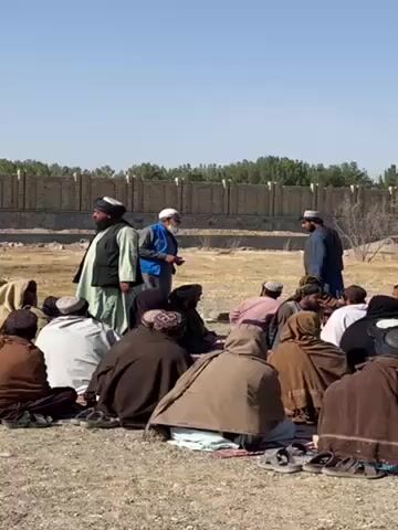 A group of men are gathered outdoors, sitting and standing in a loose circle on a dirt surface. Most of the men are wearing traditional clothing, including head coverings and robes or long coats in earth tones. Two men are standing and appear to be addressing the group. One is wearing a white beard, glasses, and a blue vest. The other is wearing a black vest over a light-colored garment. In the background, there is a low brick or stone wall with trees or bushes visible behind it. The sky is clear and blue.