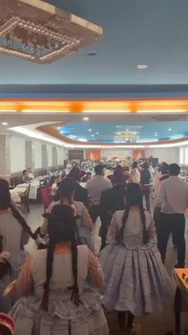 This video appears to be taken from a gathering or event inside a well-lit venue. The camera is focused on a group of women in the foreground, all wearing traditional light blue dresses with intricate white patterns and matching hats. Their hair is styled in long braids. The women are holding fans or small bags. In the background, other people are visible, seated at tables and standing, some of whom are also dressed in formal attire. The environment seems to be a banquet hall or restaurant, with long tables, chairs, and decorative lighting fixtures on the ceiling. The overall atmosphere suggests a celebratory or formal occasion.