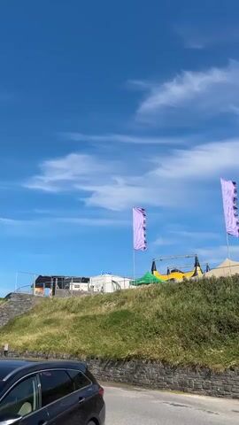 The video pans across a grassy hill under a bright blue sky with wispy clouds. Three tall, purple flags with a scalloped edge are prominently displayed, fluttering in the wind. Behind the flags, in the middle distance, is a festival-like setup with tents and what appears to be a stage or inflatable structure. A dark-colored car is partially visible in the lower left foreground. The overall scene suggests an outdoor event or festival taking place on a clear day.