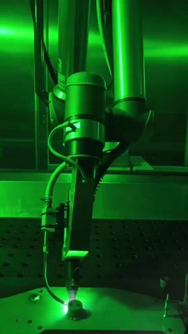 The video displays a close-up view of a high-power laser welding or cutting apparatus operating in a well-lit, industrial setting. The scene is dominated by a vibrant green light emitted from the laser, which is directed downwards onto a metal workpiece. The laser head, a complex piece of machinery with various components and tubes, is mounted on a robotic arm. A narrow tube, likely supplying gas, is connected to the welding head.

The workpiece appears to be a small, circular metal object secured to a grid-like surface, which is part of a larger workstation. The laser beam is intensely focused, creating a bright point of light and visible sparks or molten metal being ejected from the weld point. The surrounding environment is dimly lit, with the green laser light creating a striking visual effect and casting shadows. The overall impression is one of precision, power, and advanced manufacturing technology.

There is visible text on the side of the welding head, which reads "OH-au2-man". The setting appears to be a controlled environment, possibly a laboratory or a production facility, given the specialized equipment and the absence of extraneous elements. The time of day is not discernible, and there are no people or animals visible in the frame. The focus is entirely on the intricate process of laser material processing. The location cues are limited to the German context provided.