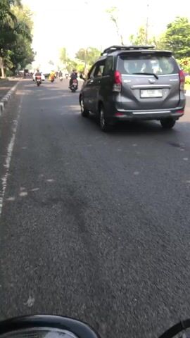 The video shows a street scene, likely in a tropical or subtropical region, with a paved road and trees lining the sides. The perspective is from a vehicle, possibly a motorcycle, as indicated by the handlebars visible at the bottom of the frame. Several vehicles are on the road: a gray SUV is in the foreground, followed by a motorcycle with a rider, and another vehicle, a black sedan, is on the right side of the road. The sun is bright, casting shadows on the road, suggesting it is daytime. The traffic appears to be moving. The background includes buildings, utility poles, and more trees. The overall impression is of a moderately busy urban or suburban street.