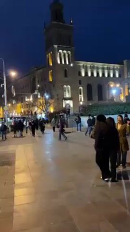 The video shows a street scene at night. Pedestrians walk on a tiled walkway in front of a large, illuminated building with a tower. A street performer stands on a raised platform, with a microphone in front of him. A man in a dark uniform stands behind him on the platform. Two people stand near the base of the platform and appear to be preparing to perform. Another person with curly hair stands between the camera and the performers, looking at the people on the platform.