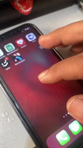 A pair of hands is shown interacting with a smartphone. The phone is black with a screen that displays a red wallpaper with a striped pattern. An icon for an app labeled "Work Zone" is visible. The finger then taps on the screen, and the phone's display changes to a black screen with a white progress bar. The screen then transitions to a bright yellow background with a loading indicator and a small square icon. The camera is positioned above and to the side of the phone, focusing on the screen and the hands.