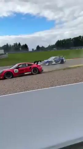 The video shows race cars passing by on a track. The person recording is standing near a chain-link fence, capturing the cars as they speed past. A red and pink car with the number 84 and a yellow car are visible. In the background, there's an excavator, grass, trees, and a cloudy sky. Later, the camera pans to show an electric scooter parked near the fence next to a road case.