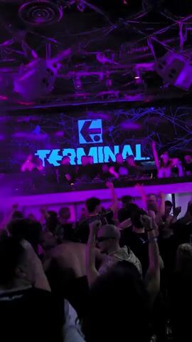 Here is a description of the video:

The video captures a vibrant nightlife scene within a club or concert venue. In the foreground, a crowd of people are seen dancing and raising their arms in the air. They are silhouetted against the bright stage, indicating a focus on the performers. 

The stage itself is elevated and crowded with individuals who seem to be performers or DJs. Behind them, a large screen displays a logo with the word "TERMINAL" prominently featured. The lighting in the venue is dynamic, with blue and purple hues illuminating the stage and audience. The overall atmosphere is energetic and lively, typical of a music event.