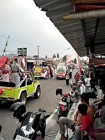 The video captures a street scene, possibly a parade or festival, in what appears to be a town or village. A bright green open-top vehicle, resembling a modified Jeep, is driving down the street from left to right. It is followed by a white truck decorated with various colorful elements, including what looks like a large circular painting or display in the center, and is being propelled by an unseen mechanism. There are people lining the sides of the street, some seated on benches and others standing. Many of them are holding or wearing red and white elements, suggesting a patriotic or celebratory event. Several individuals in traditional or festive attire are walking alongside or behind the vehicles. There are also motorcycles parked on the side of the road. Buildings with metal roofs and some shops are visible on the right side of the street. Indonesian flags are prominently displayed on poles and draped across some structures. The sky is overcast, and the overall atmosphere suggests a public gathering.