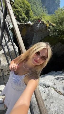The video opens with a close-up selfie of a young woman with blonde hair, wearing sunglasses, a white dress, and a pearl necklace, smiling at the camera. She is standing on a wooden walkway with metal railings, with a rocky, mountainous landscape and lush green trees in the background. A waterfall is visible to her left, cascading down into a deep gorge. The camera then pans to show another person, possibly a man, in the distance taking a selfie on the same walkway. The video then shifts focus to the waterfall and the gorge, showing the powerful flow of water and the sheer rock faces of the canyon. In the distance, more people can be seen near the edge of the gorge. The overall scene depicts a natural landscape with a waterfall and canyon, with people enjoying the scenery and taking photos.