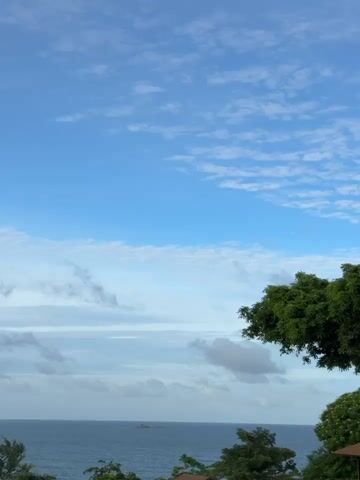 The video shows a wide shot of a tropical seascape. The sky is a bright blue with scattered white clouds, some of which are wispy and others are more substantial. The ocean is a deep blue, meeting the horizon where a small island or rock formation is visible in the distance. In the foreground, lush green trees frame the right side of the scene, with their leaves rustling gently in what appears to be a light breeze. The overall impression is one of a peaceful, natural landscape, likely taken from an elevated vantage point. There are no people or man-made objects visible apart from a distant vessel on the water.