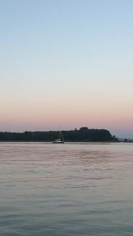 The video shows a calm body of water, possibly a lake or bay, during sunset. The sky transitions from a pale blue at the top to soft pink and purple hues near the horizon. A sailboat is visible in the distance on the left side of the frame, its sail white against the muted colors of the sky. The water is tranquil with gentle ripples. On the right side of the frame, a floating blue dock with a ladder is present. In the far distance, silhouetted mountains are visible along the horizon. The overall atmosphere is serene and peaceful.