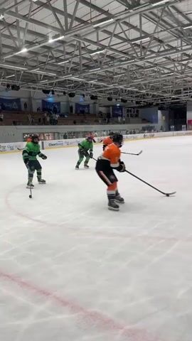 A group of people in hockey gear are on an ice rink. Two players in green and orange uniforms are skating near the center of the rink, engaged in a hockey game. Other players in similar uniforms are scattered across the ice. The rink is enclosed by a glass barrier and netting, with spectators visible in the background. The ceiling of the arena is visible, with exposed beams and lighting fixtures. A scoreboard is displayed on the wall in the distance. The overall scene depicts a recreational or amateur hockey game in progress.