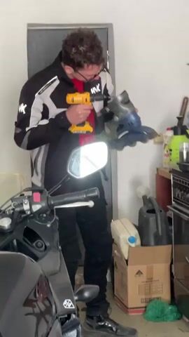 A man is using a yellow power drill to work on a pair of roller blades in a garage. He is wearing a black and white motorcycle jacket, black pants, and black shoes. He also has glasses and short, curly hair. He is standing next to a black motorcycle. Behind him is a closed door, a cardboard box, a watering can, and cleaning supplies on a shelf.