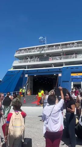 The video shows a busy port with a large ferry docked and a group of people disembarking or preparing to board. The ferry, painted blue and white with a Greek flag prominently displayed, has its ramp lowered, allowing access to the vehicle deck. Several people in bright orange vests appear to be port staff or crew, guiding the passengers. The passengers are diverse, carrying luggage, and forming queues. In the background, several white buildings and buses suggest a transportation hub. The weather appears sunny and clear with a bright blue sky. The video then transitions to a wider shot of the port, showing more people and buses, reinforcing the sense of a busy travel day.