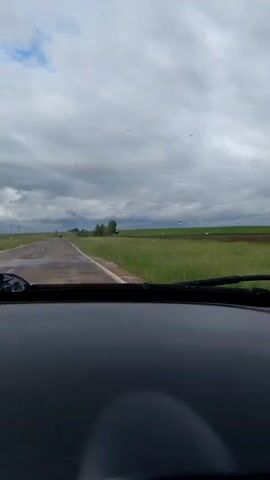 The video shows a view from inside a car, looking out through the windshield. The car is driving on a paved road that stretches into the distance. On either side of the road, there are fields of green grass. The sky is overcast with clouds, and there are some patches of blue sky visible. There are a few raindrops on the windshield. In the distance, there are some trees and what appear to be power lines. The video is shot at a slight downward angle, showing the dashboard of the car in the foreground.